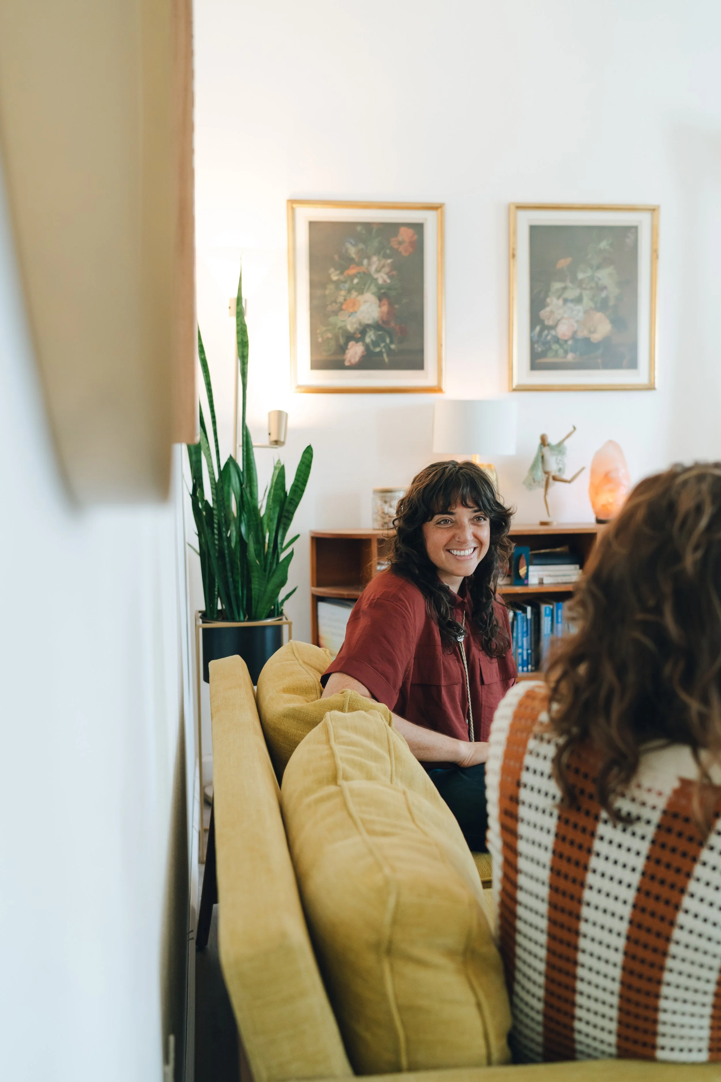 A woman with curly brown hair wearing a red shirt, sitting on a yellow couch and smiling.