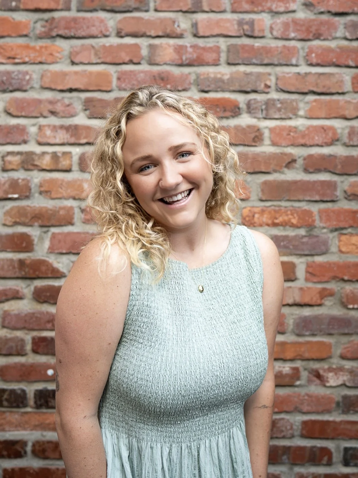 A smiling young woman with curly blonde hair wearing a sleeveless light green dress standing in front of a brick wall.