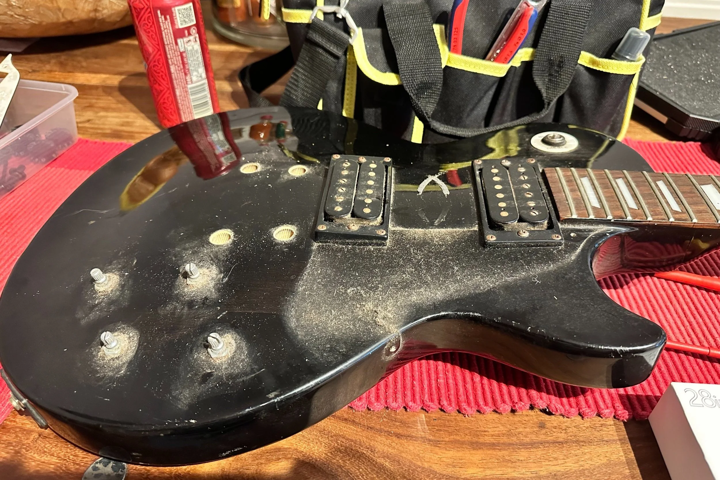 A black electric guitar with no strings, showing dust and bolt holes, resting on a red cloth on a wooden surface. In the background, there is a black tool bag, a red object, and various small parts.