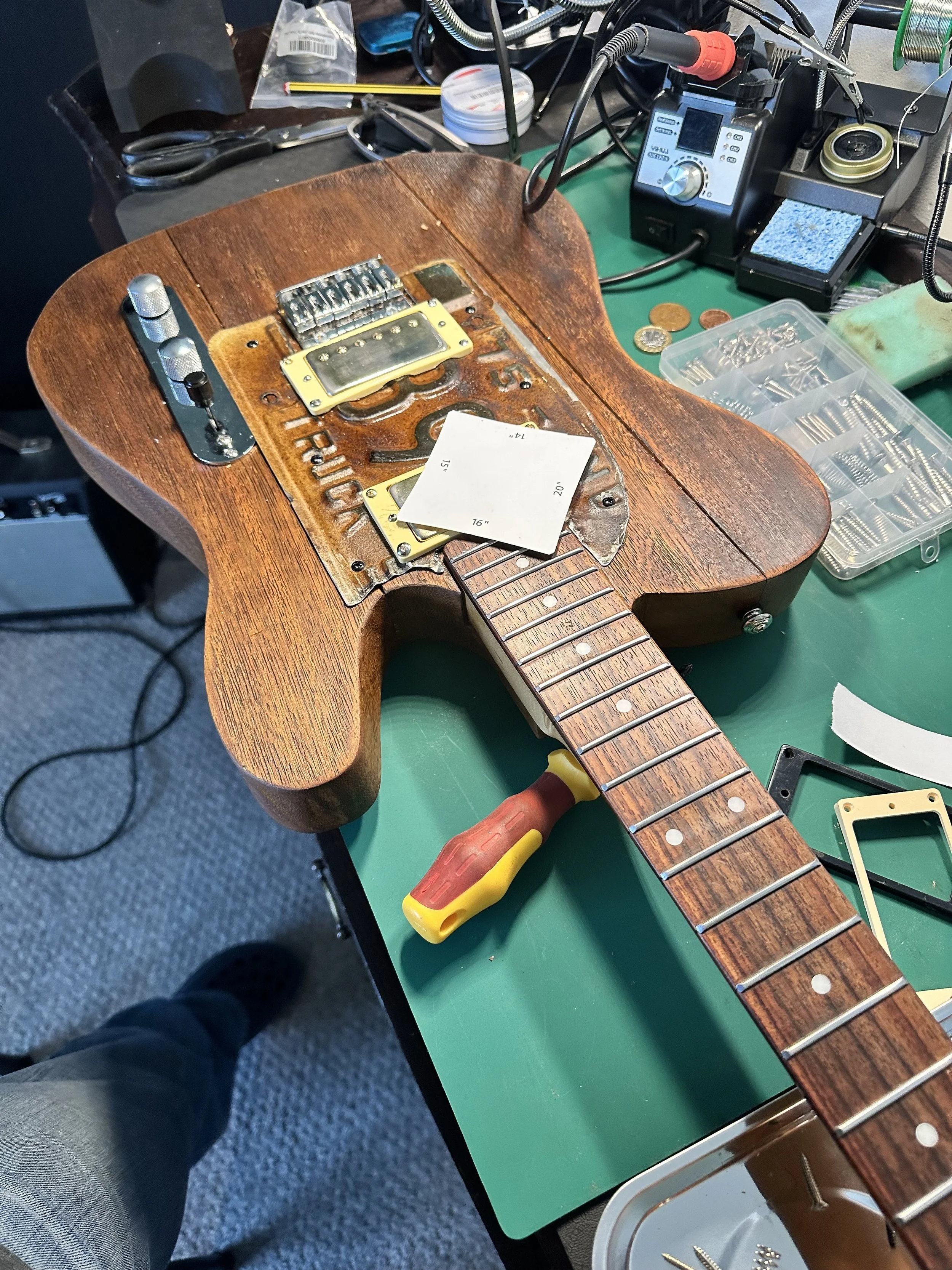 A partially disassembled electric guitar on a green workbench, with tools, screws, and parts around it, in a workshop setting.