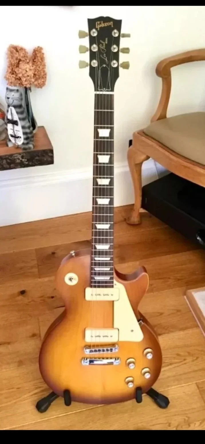 A Gibson electric guitar with a sunburst finish on a guitar stand, placed on a wooden floor near a chair and a side table with decorative items.