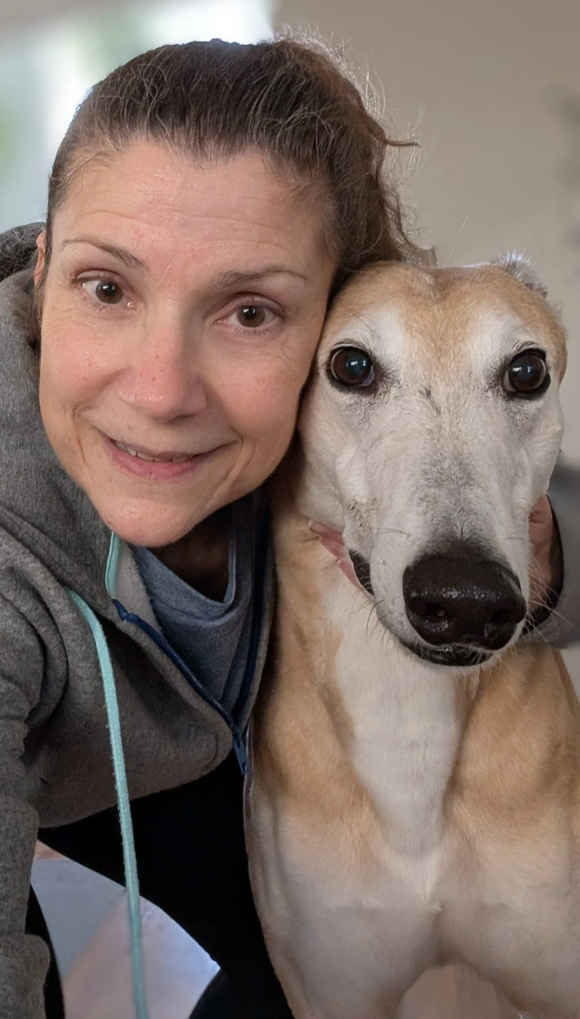 A woman with brown hair in a ponytail taking a selfie with a tan and white dog, both appear close together with their faces next to each other.