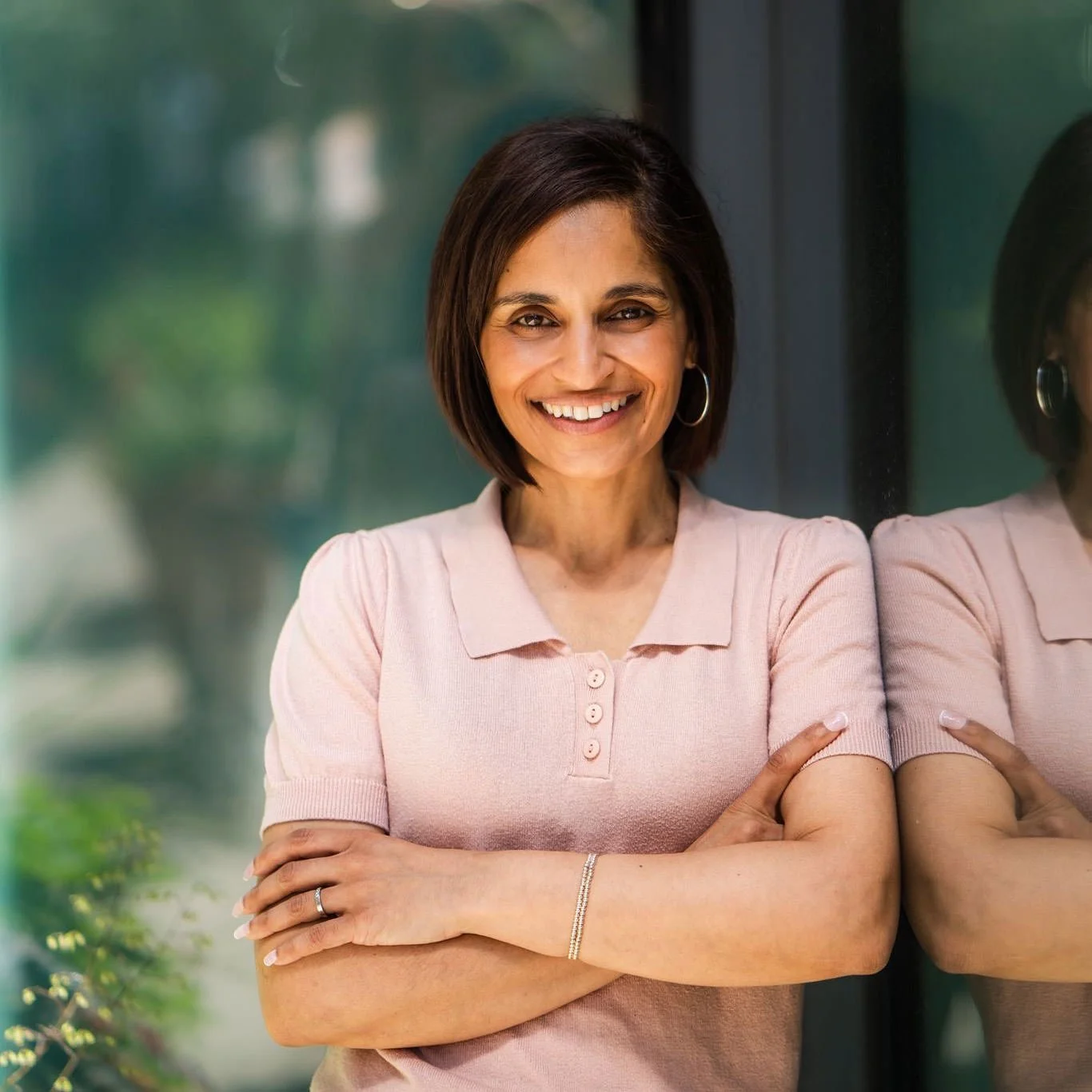 A woman with short dark hair and hoop earrings standing with arms crossed, smiling, next to a window with a reflection.