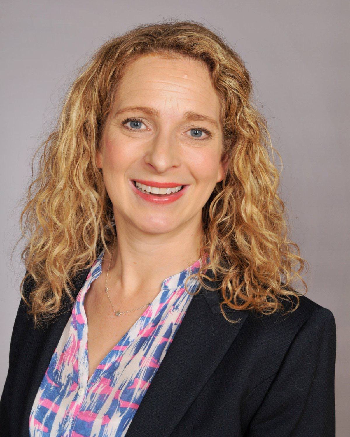 A woman with curly blonde hair, blue eyes, and light skin, smiling, wearing a black blazer and a multicolored blouse with pink, white, and blue patterns, posed against a light gray background.