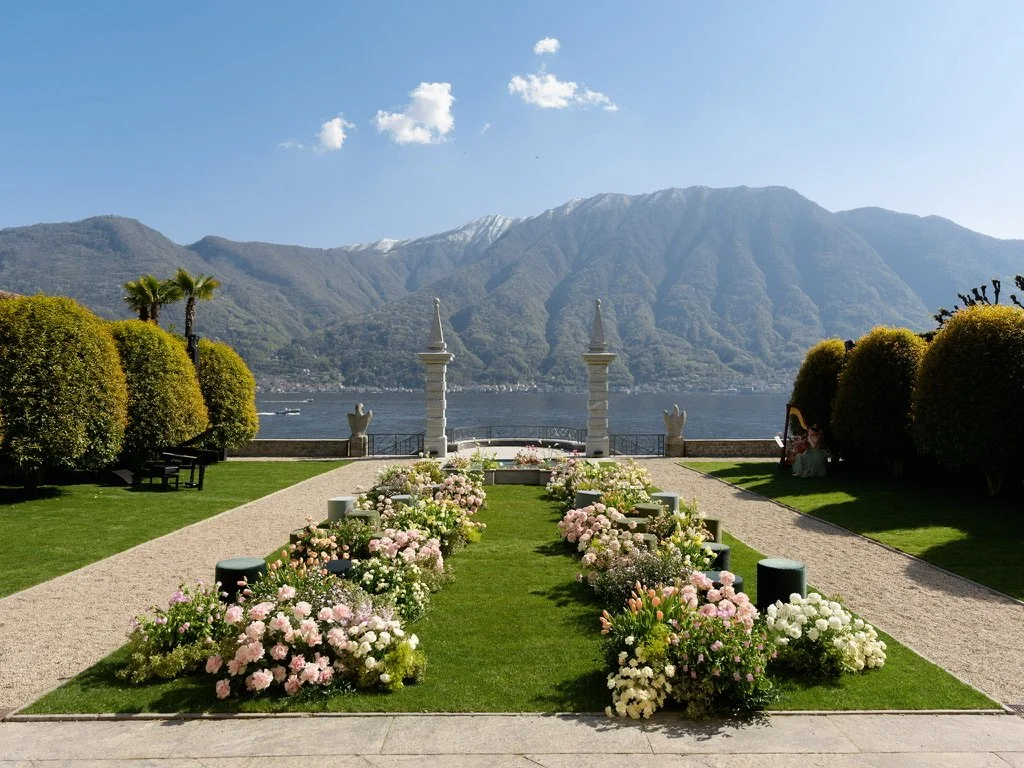Flower-lined lawn aisle with velvet poufs leading to two stone pillars and Lake Como at Villa Balbiano