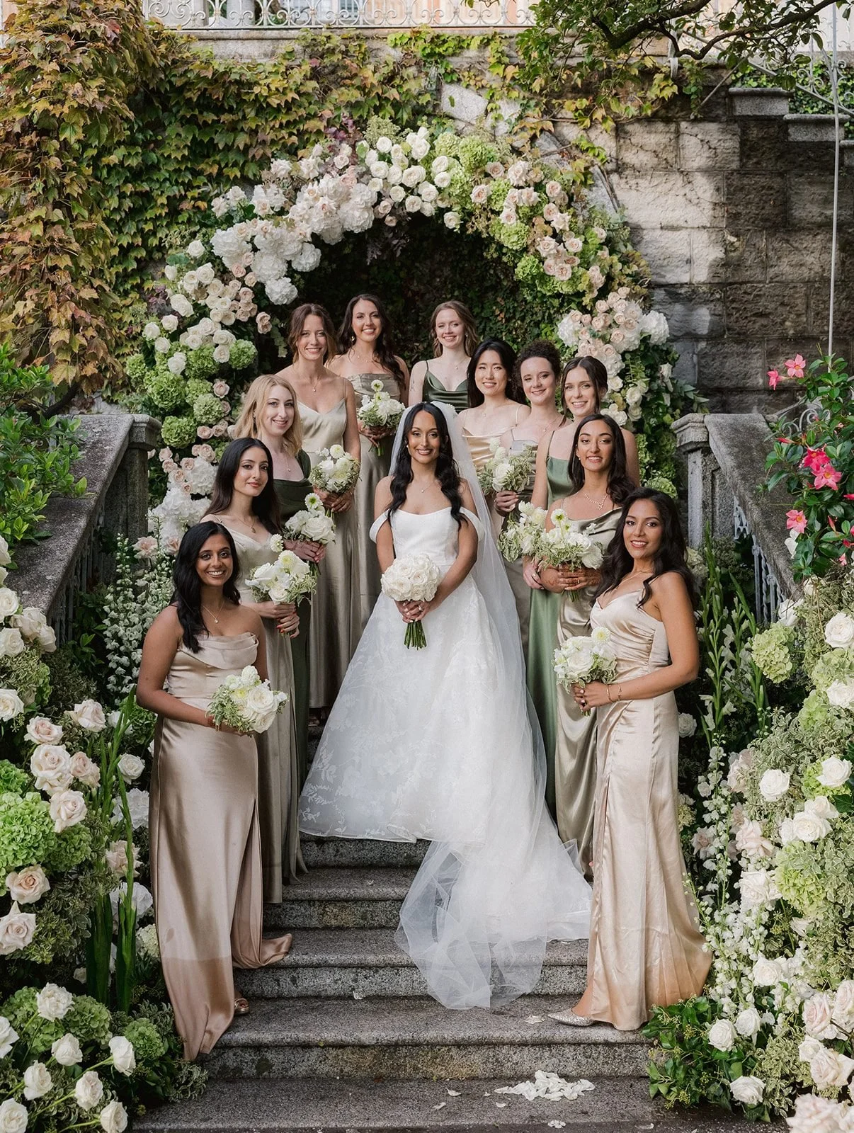 A bride and her bridesmaids in sage and champagne satin on the stone stairs of Villa Cipressi beneath an arch of white and blush roses and green hydrangea.