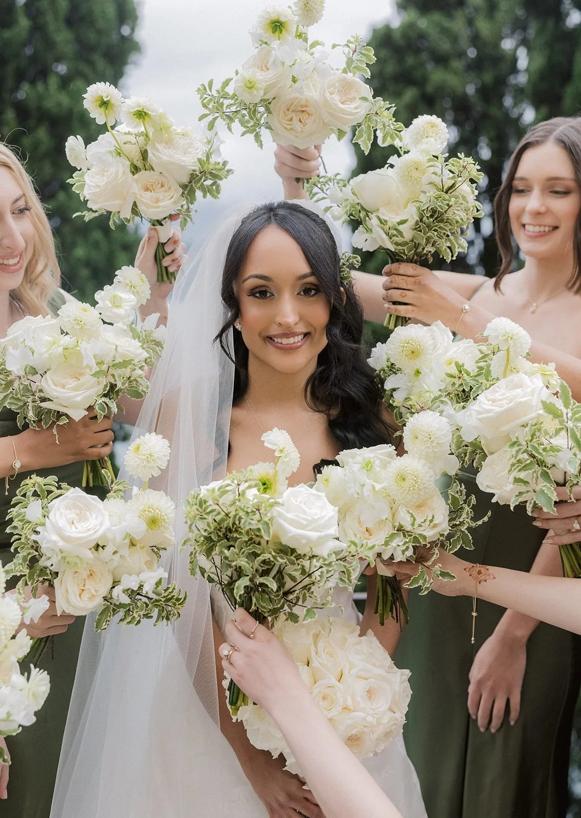 Close-up of a smiling bride holding an all-white rose bouquet, surrounded by bridesmaids' bouquets of white garden roses, white pompon dahlias and variegated greenery.