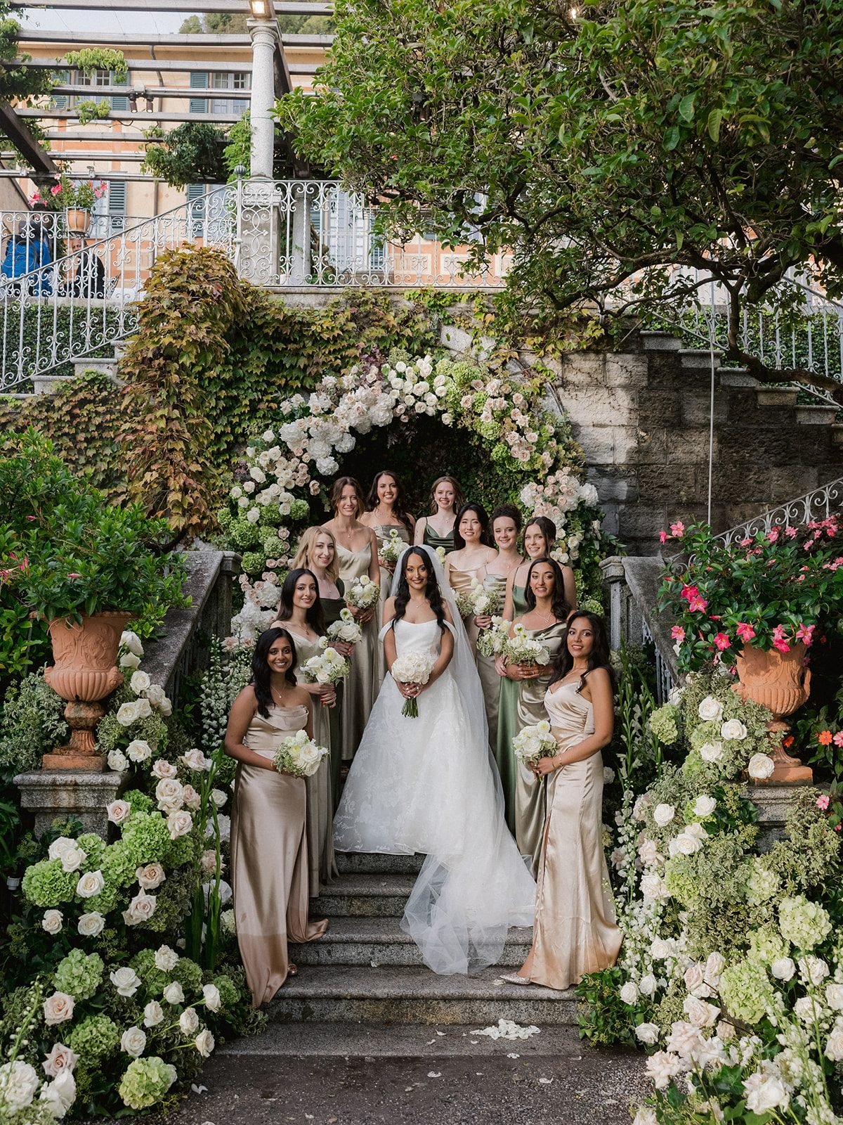 Wide view of the bridal party on Villa Cipressi's stone staircase, framed by a rose and hydrangea arch, terracotta pots and white flowers along the steps.