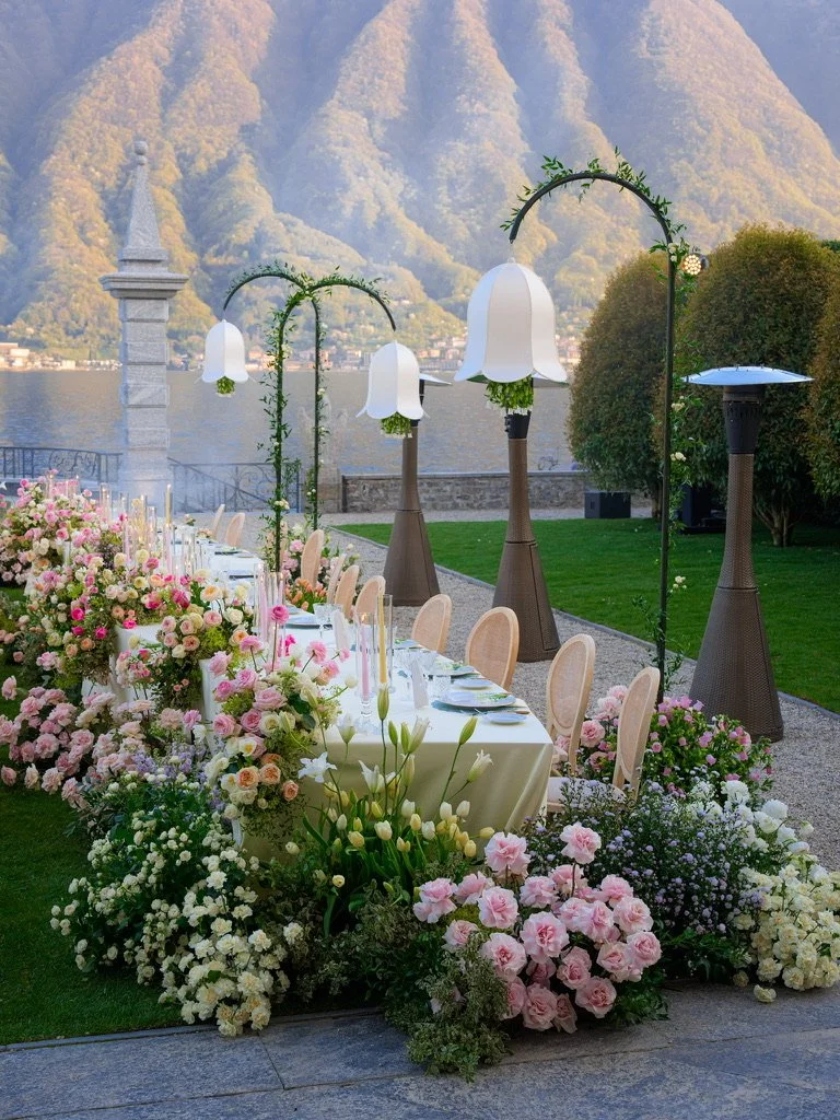 Dinner table with white bell lanterns on green arches and pastel florals spilling onto the lawn
