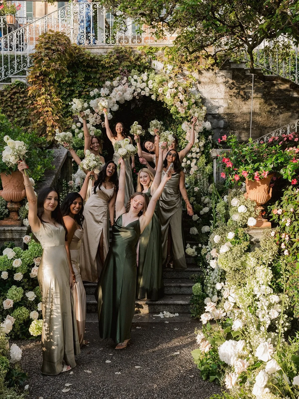 Bridesmaids in sage and champagne satin cheering with bouquets raised on the stone stairs under the rose and hydrangea arch at Villa Cipressi.