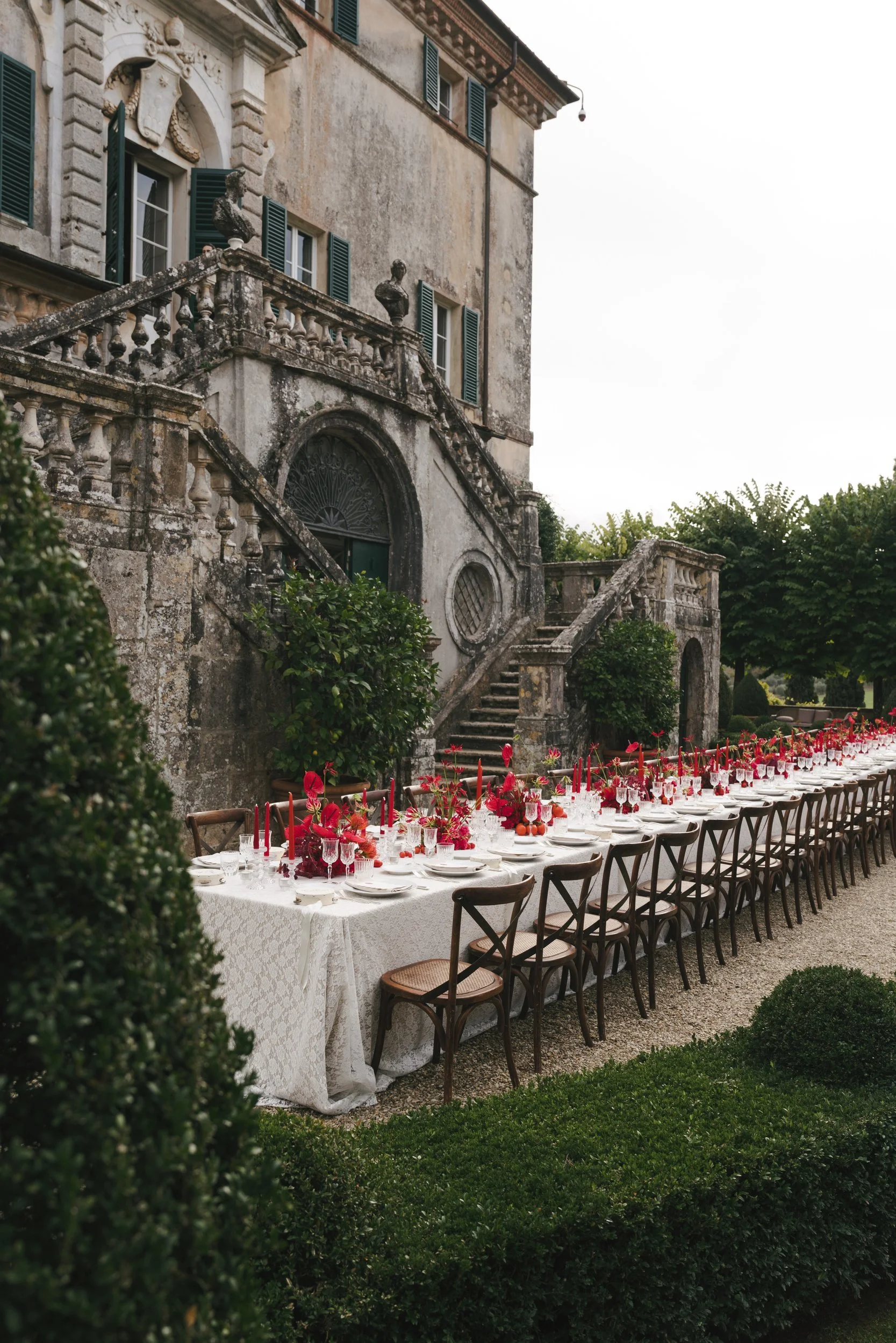 Long lace-covered welcome dinner table with deep red floral arrangements beneath the double stone staircase of Villa Cetinale, Tuscany