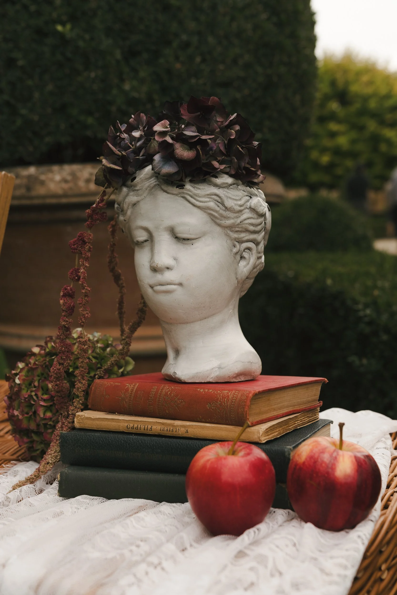 Classical bust crowned with burgundy hydrangea on antique books beside red apples and trailing amaranthus