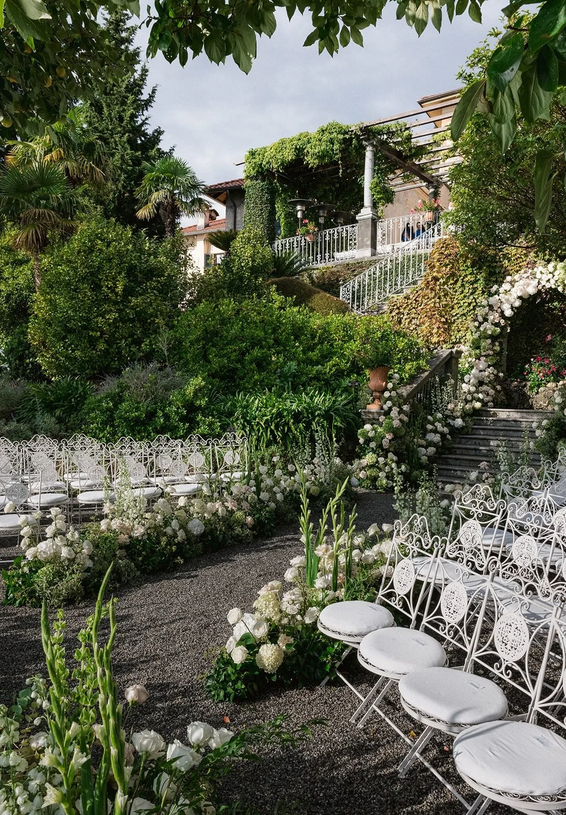 White wrought-iron chairs beside an aisle of white roses, hydrangea and gladiolus, with the terraced gardens and stone stairs of Villa Cipressi rising behind.