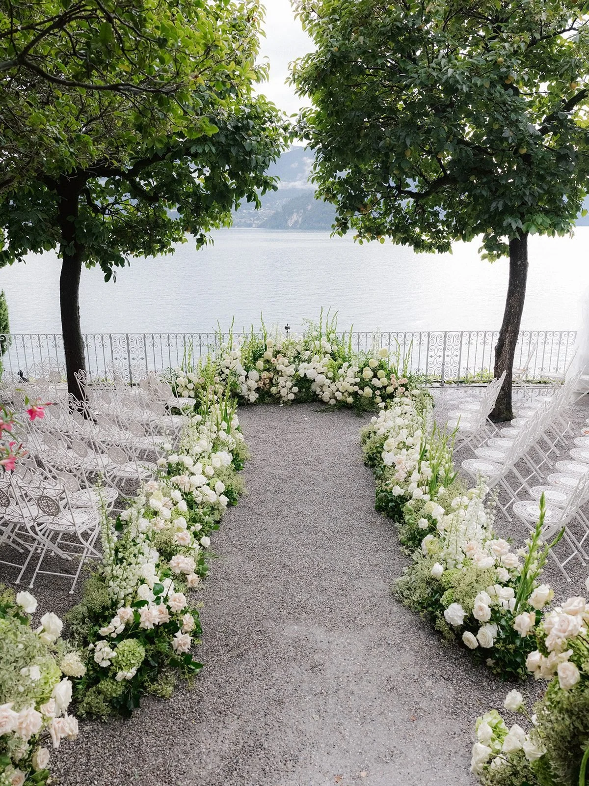 A lakeside terrace ceremony at Villa Cipressi in Varenna, with a gravel aisle lined in white roses and hydrangea, white wrought-iron chairs and Lake Como beyond the trees.