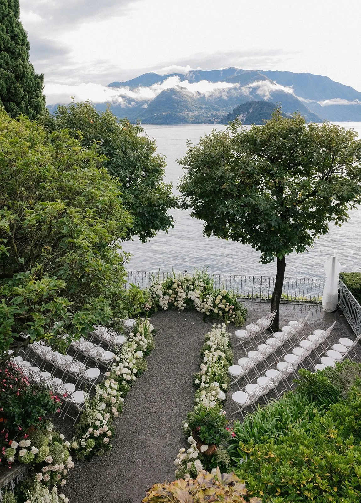 View down the Villa Cipressi gardens to a lakeside ceremony aisle edged with white flower meadows, rows of white chairs and Lake Como with mountains behind.