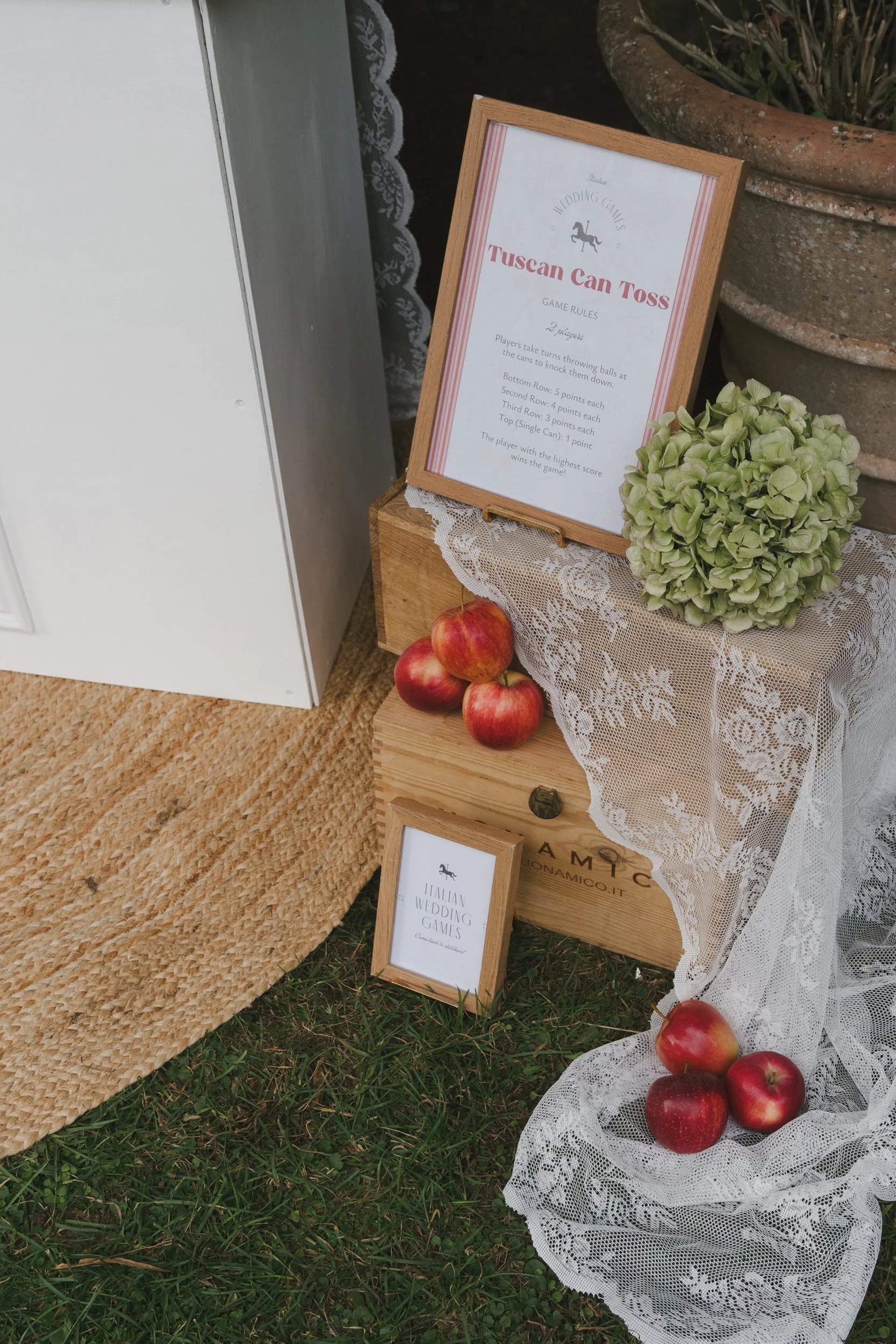 Italian wedding games sign on a wooden crate with green hydrangea, draped lace and red apples on the lawn