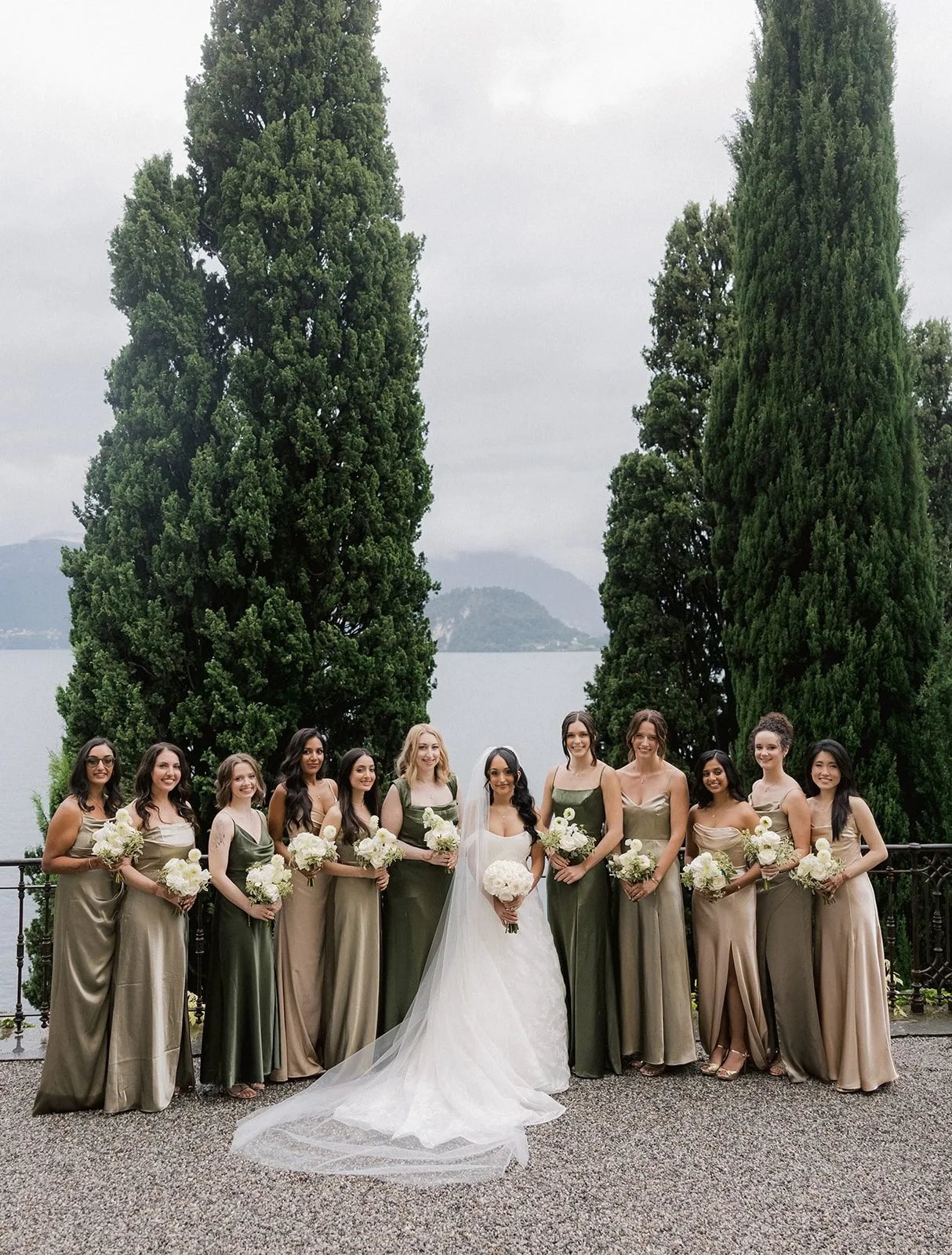 A bride and twelve bridesmaids in sage and champagne satin dresses holding white bouquets on the lakeside terrace, with tall cypress trees and Lake Como behind.