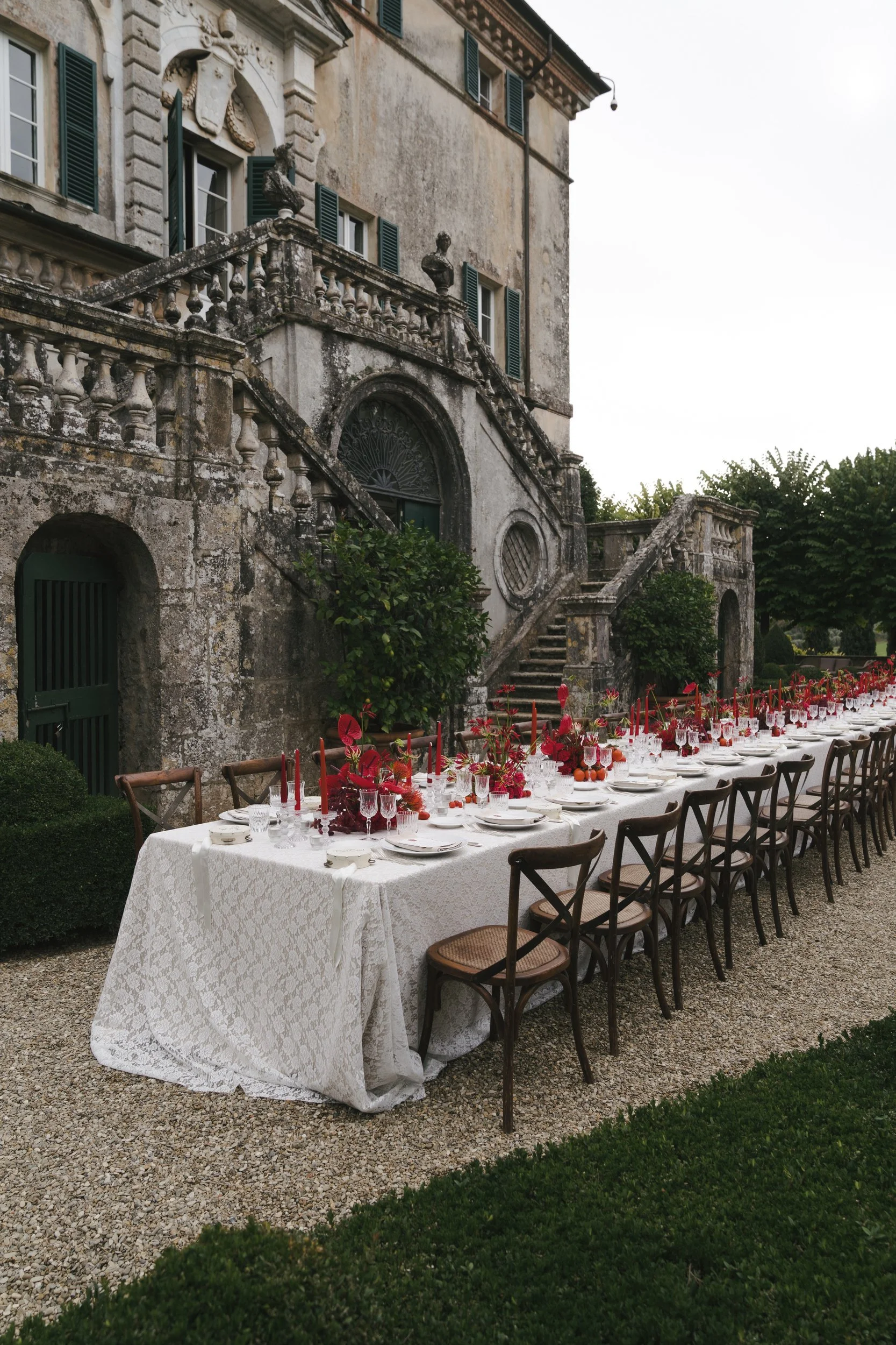 Wide view of a long Tuscan welcome dinner table with cross-back chairs and red centrepieces in front of Villa Cetinale's baroque facade