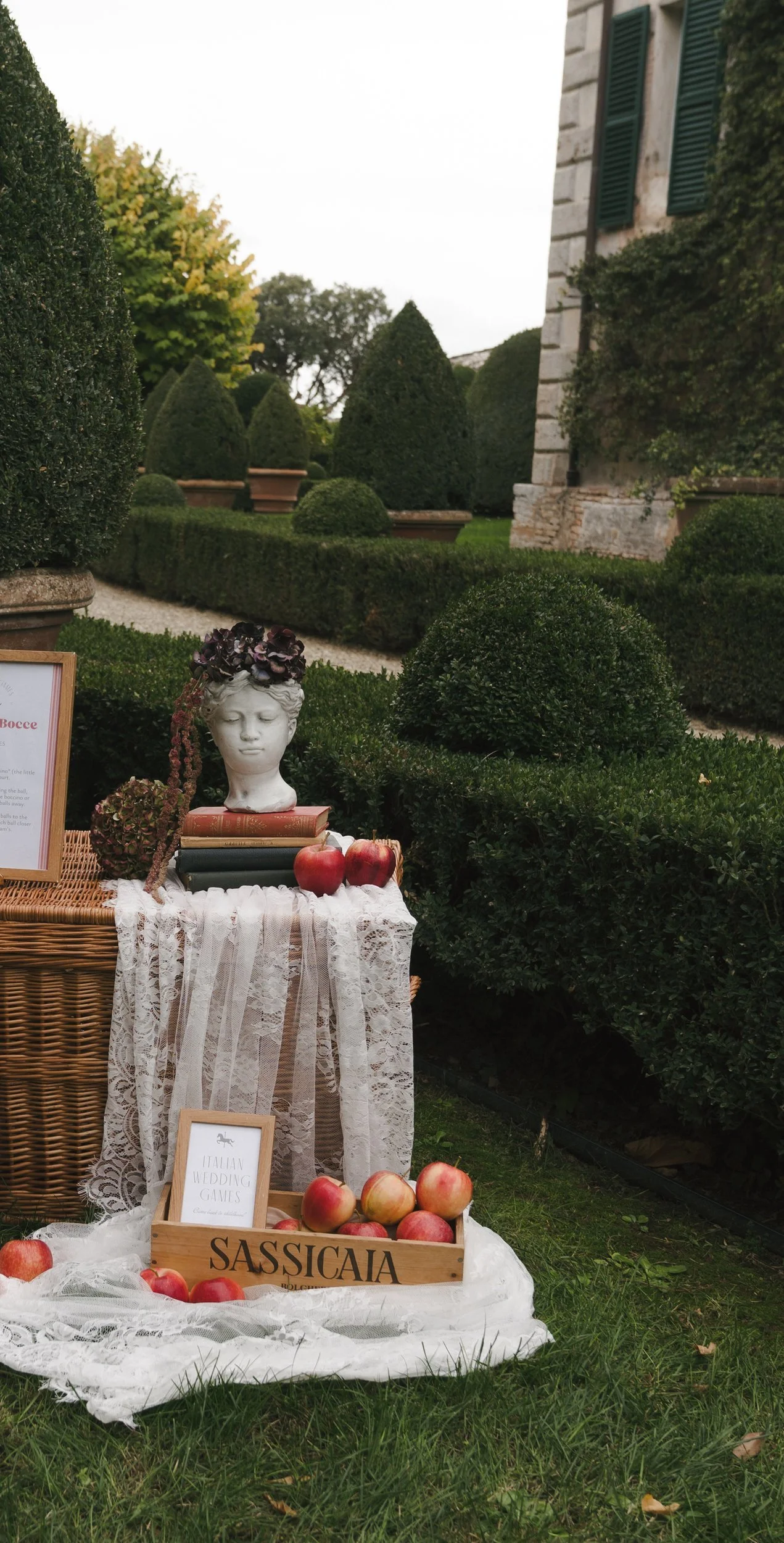 Garden vignette with a classical bust, antique books, wicker hamper, lace and red apples at Villa Cetinale