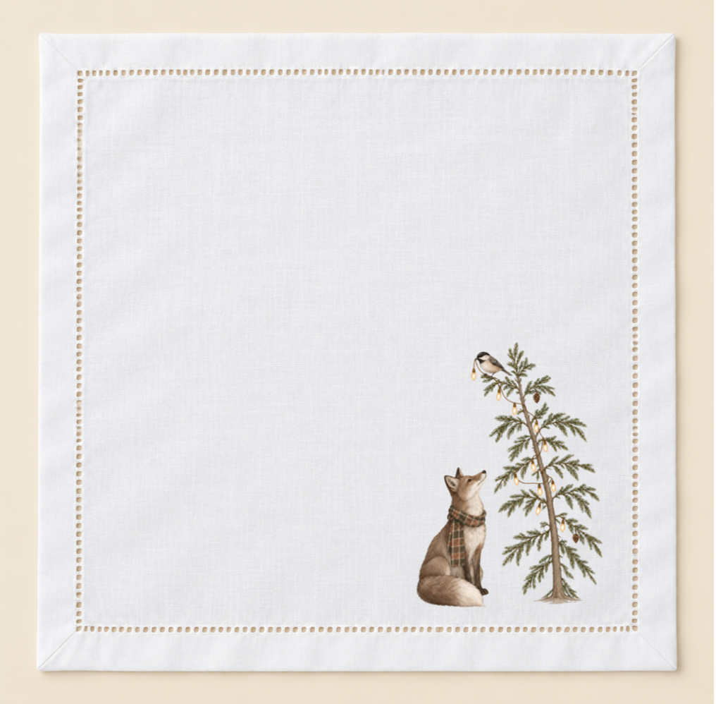 White 20-inch hemstitched cotton napkin with a hand-pressed woodland design featuring a fox in a plaid scarf beside a lit evergreen tree and chickadee.