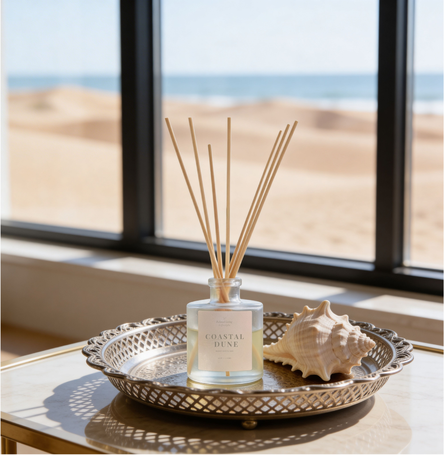 Coastal Dune reed diffuser displayed on textured metal tray with white shell, coastal home fragrance with ocean and sand dunes view through large windows.