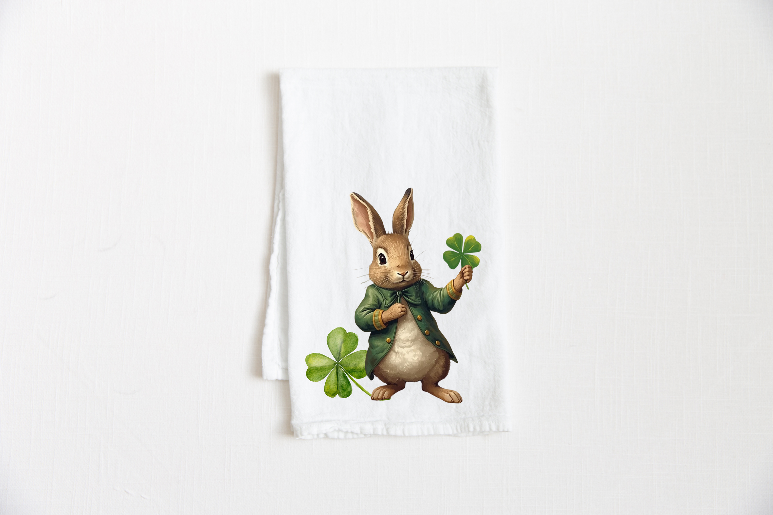 St. Patrick’s Day rabbit tea towel in flat lay on white background featuring a bunny in a green coat holding a four leaf clover, 100% cotton 27x27 spring kitchen decor.