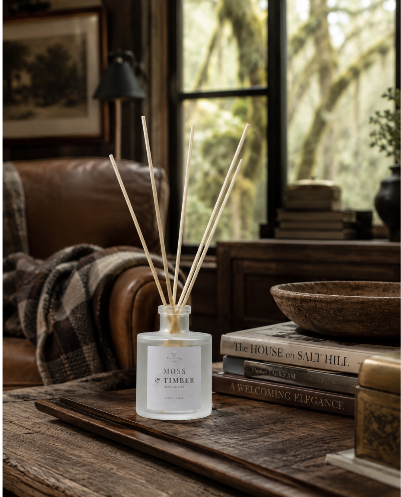 Moss & Timber reed diffuser styled in a wooden tray with decorative books in an elevated nature-inspired living room, featuring a blurred view of moss-covered trees outside the window.