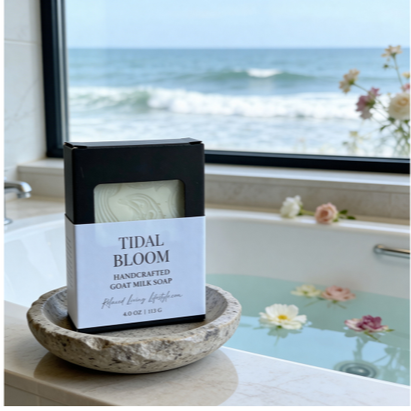 Tidal Bloom goat milk soap bar styled beside a soaking tub with subtly scattered floating flowers in the water, set near a large window with a soft ocean view in the background.