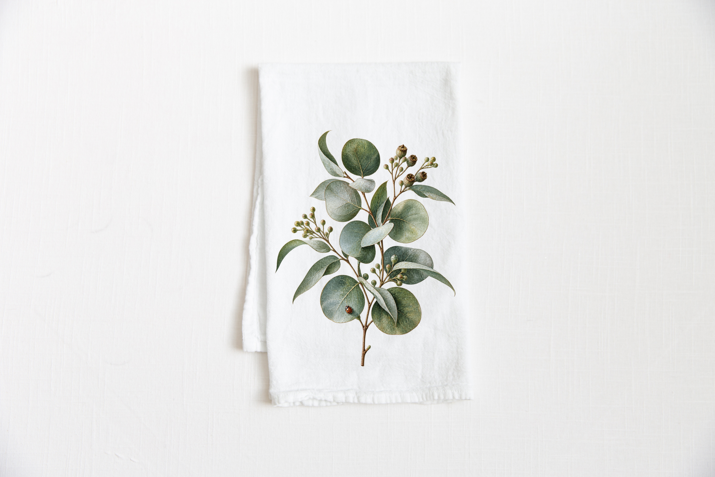 Eucalyptus Cotton Tea Towel featuring soft silver-green eucalyptus leaves, seeded stems, and a small ladybug on a white tea towel