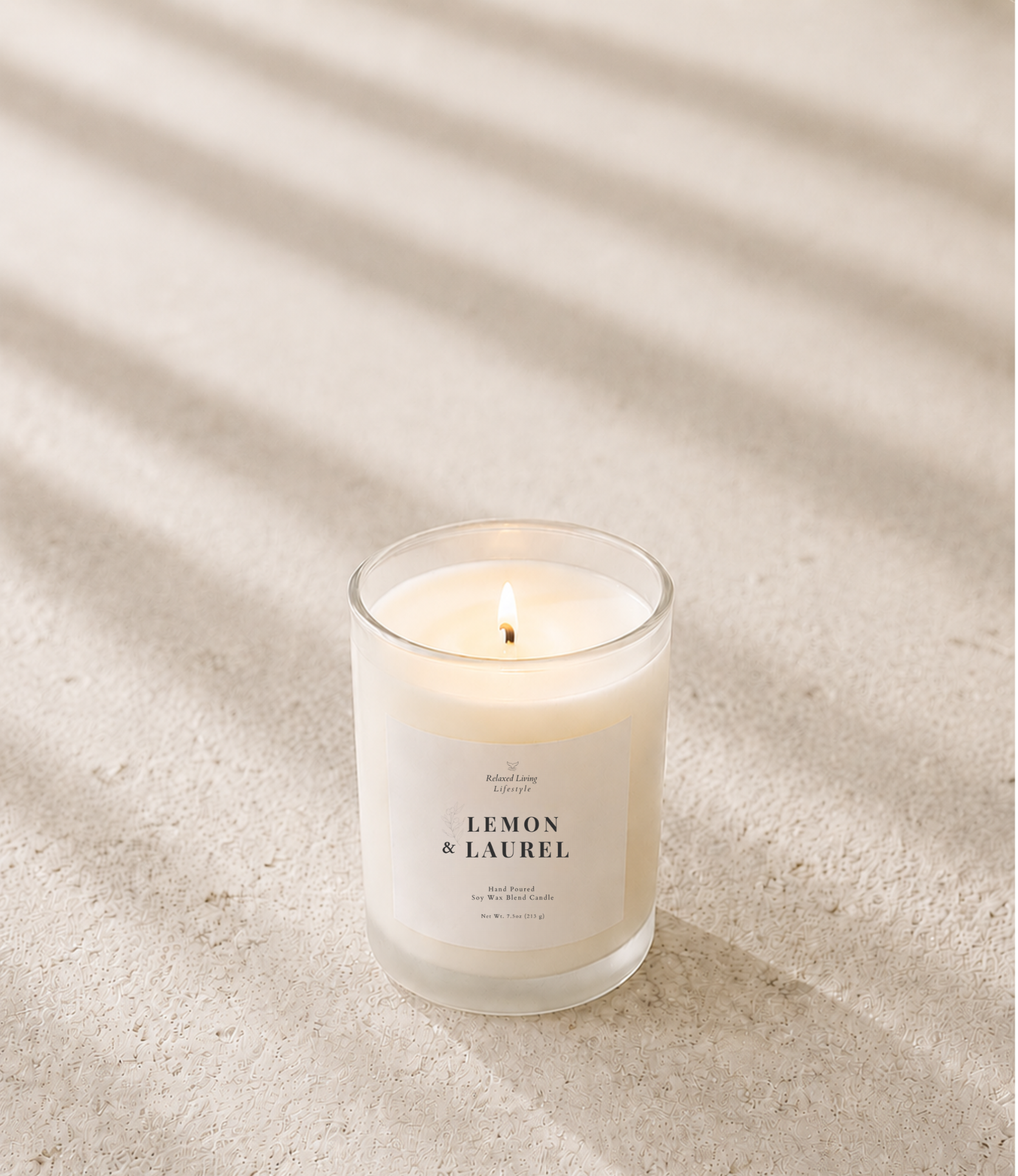 Lit frosted glass soy wax candle on a light ivory travertine surface with soft natural window light and subtle diagonal shadows.