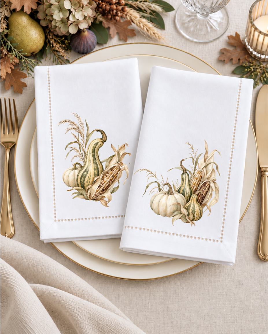 Two white hemstitched napkins with coordinating fall botanical designs arranged on a neutral place setting with gold flatware and autumn accents.