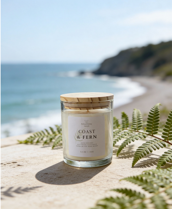 Coast & Fern 5 oz soy candle styled with airy ferns on textured surface, coastal view in background. Bergamot, lavender, moss, and juniper scent.