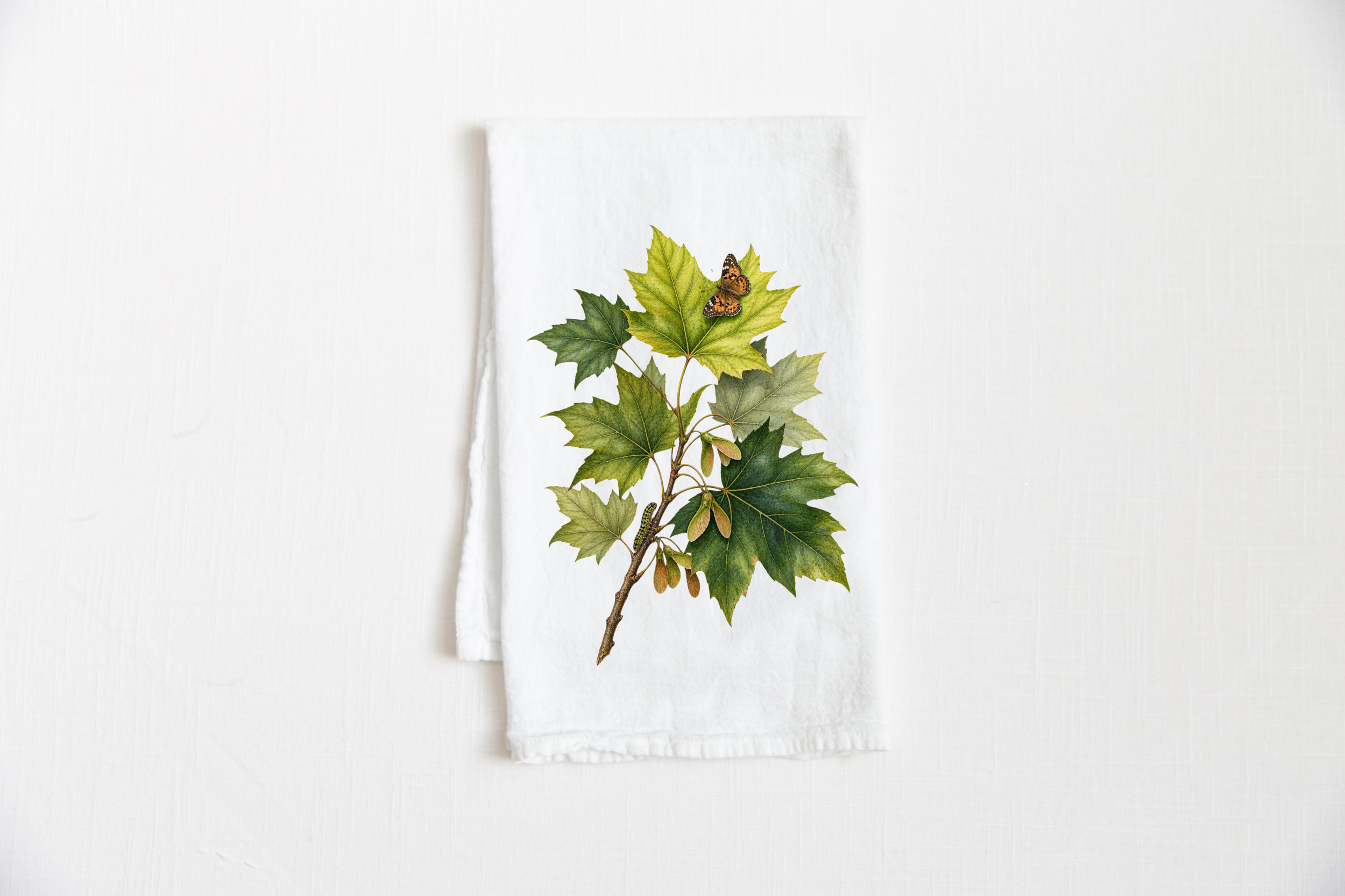 White cotton tea towel featuring a botanical illustration of green and golden autumn leaves with a caterpillar and butterfly.