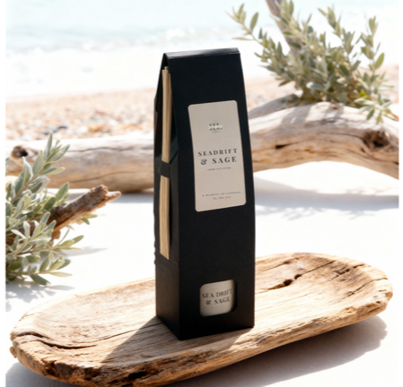 “Seadrift & Sage boxed reed diffuser displayed on a wood tray at the beach with driftwood and ocean waves in the background, coastal-inspired home fragrance ideal for relaxing, nature-inspired décor.”