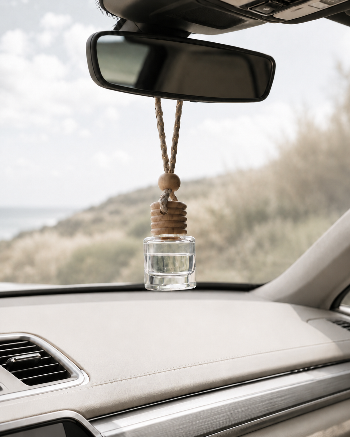 Sea Drift & Sage car diffuser with a light wood cap and clear oil hanging from a rearview mirror inside a neutral car interior, with softly blurred sage-toned grasses and greenery outside