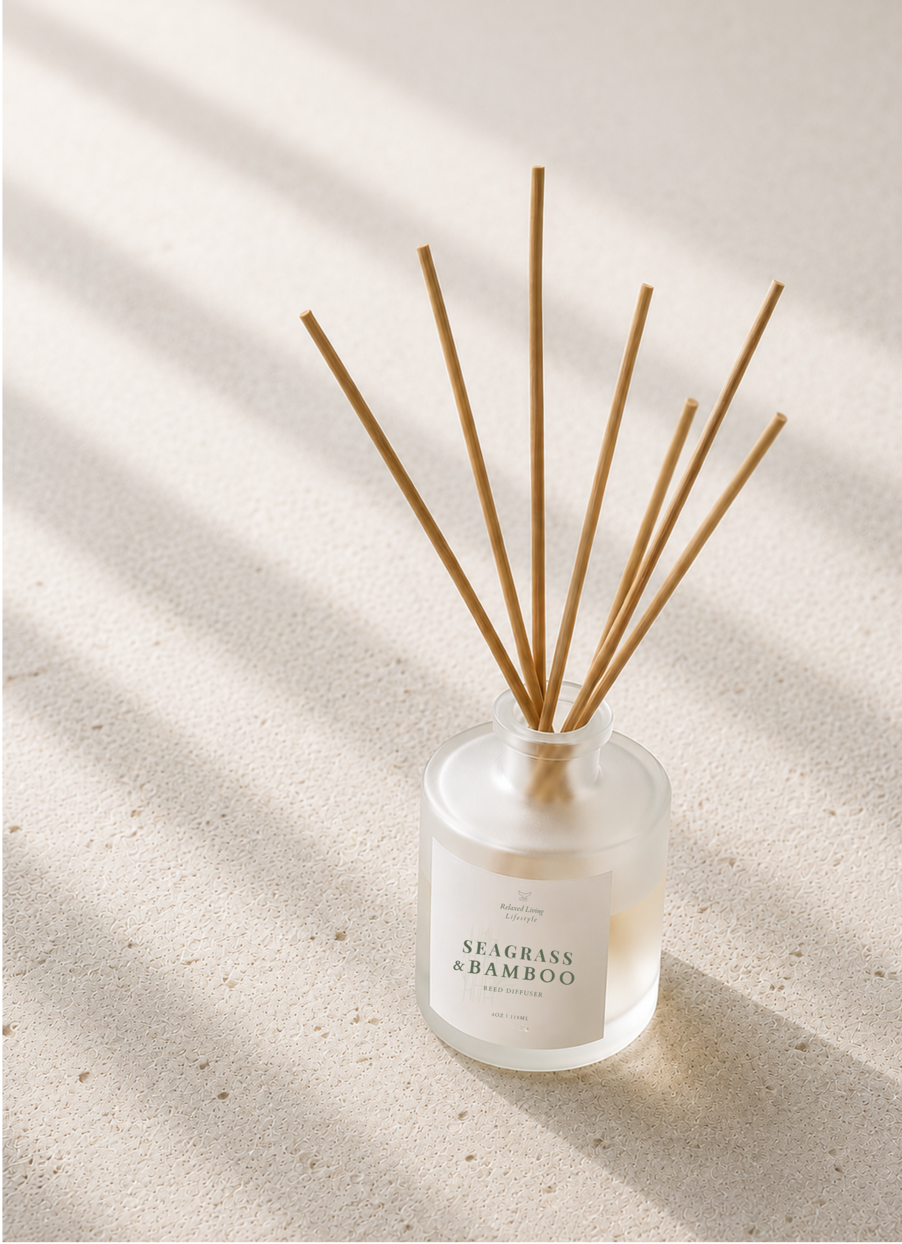 Frosted glass reed diffuser with natural rattan reeds on a light ivory travertine surface illuminated by soft afternoon window light.