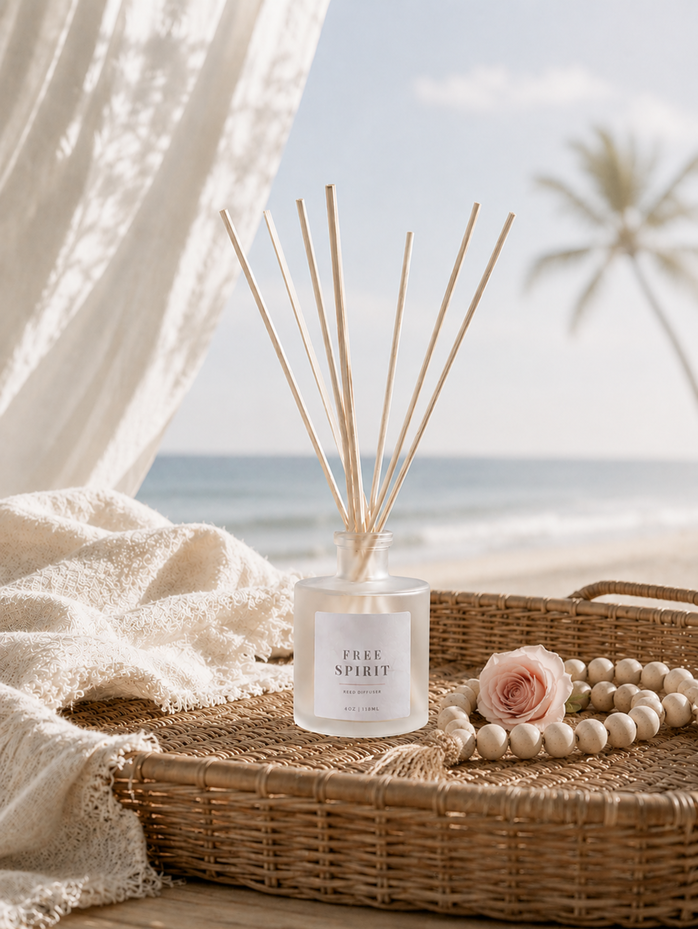 Free Spirit reed diffuser styled on a rattan tray with soft rose accents and clay beads beside the diffuser, overlooking a blurred ocean view for a relaxed luxury coastal home fragrance aesthetic.