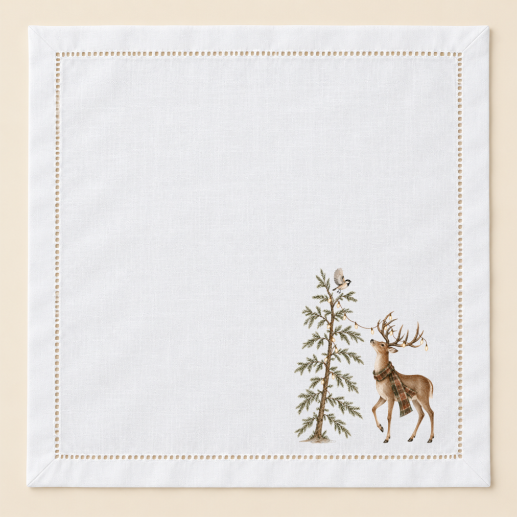 White 20-inch hemstitched cotton napkin with a coordinating hand-pressed woodland design featuring a deer in a plaid scarf beside a lit evergreen tree and chickadee.