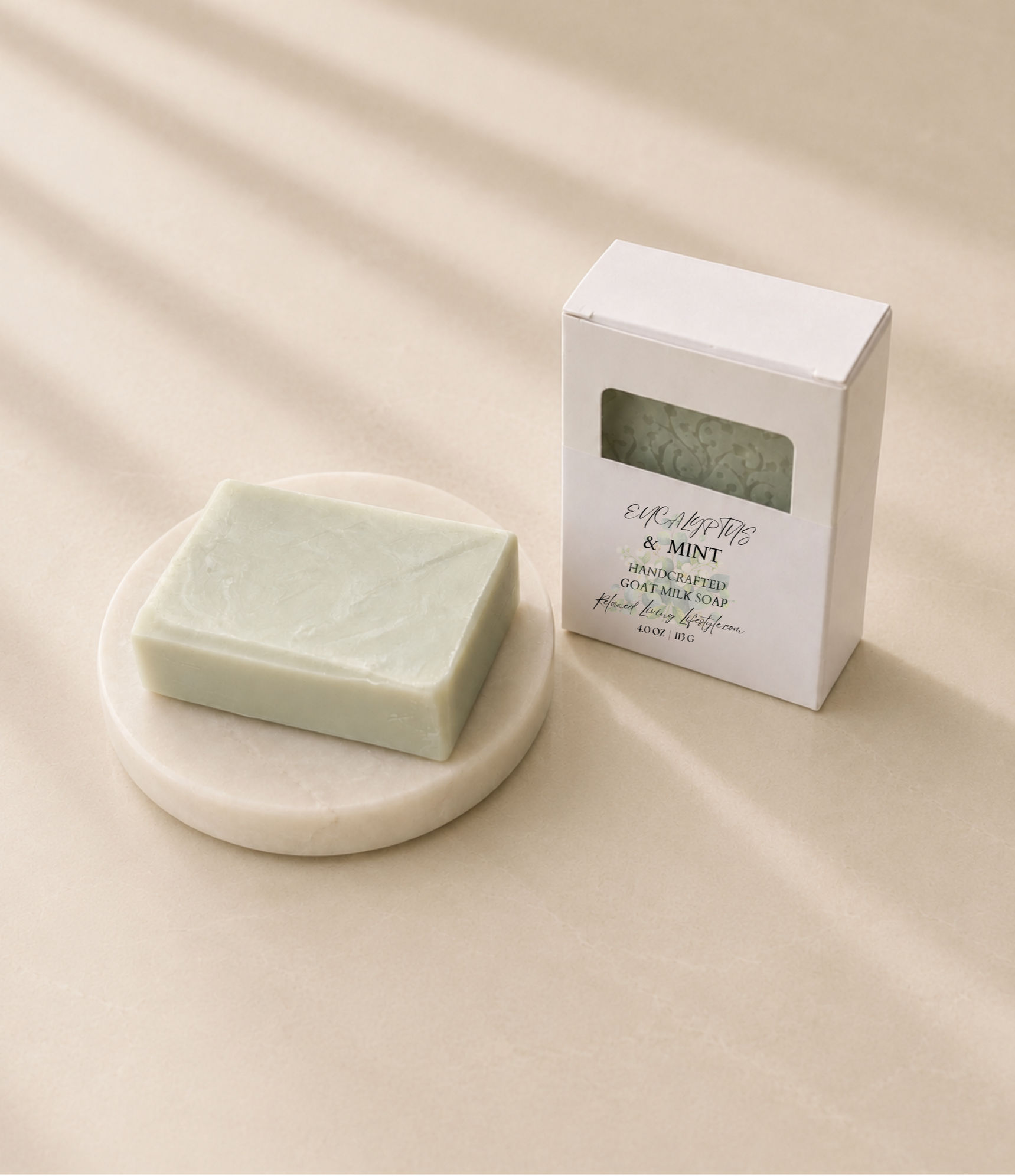 Eucalyptus & Mint handcrafted goat milk soap on a round marble soap dish beside its retail box on a smooth honed limestone surface with soft afternoon sunlight and elegant diagonal window shadows