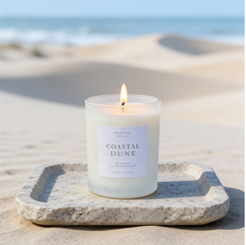 Coastal Dune soy candle lit on a textured stone tray, set against a subtle view of sand dunes and ocean, creating a soft coastal atmosphere with a clean, elevated aesthetic