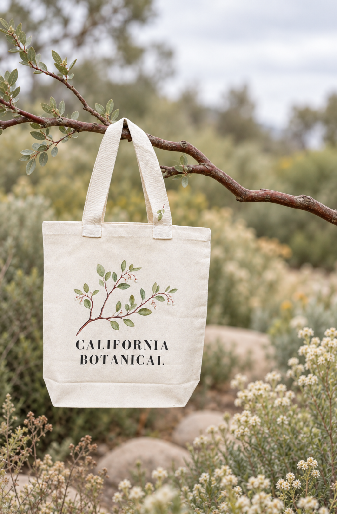 California Botanical Canvas Tote Bag with botanical branch artwork and leaf-print handles, hanging from a tree branch in a wildflower garden