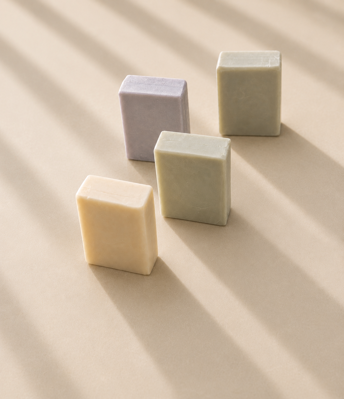 Four handcrafted goat milk soap bars displayed on a smooth honed limestone surface with soft afternoon sunlight and elegant diagonal window shadows.