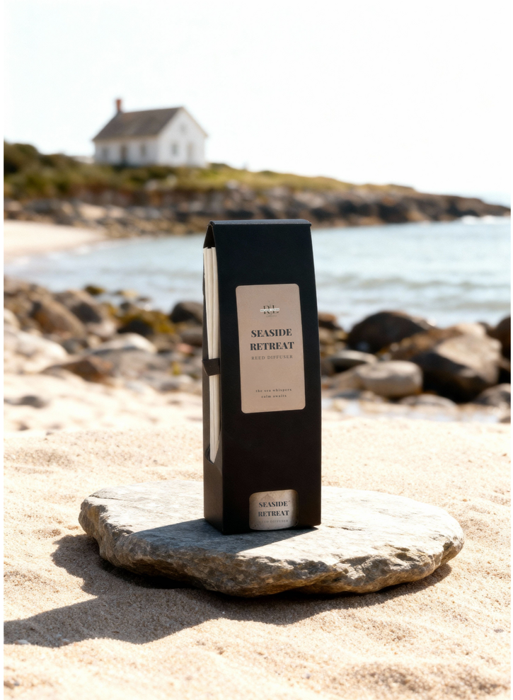 “Seaside Retreat reed diffuser and box displayed in front of a coastal cottage setting on the, evoking serene ocean-inspired home fragrance.”