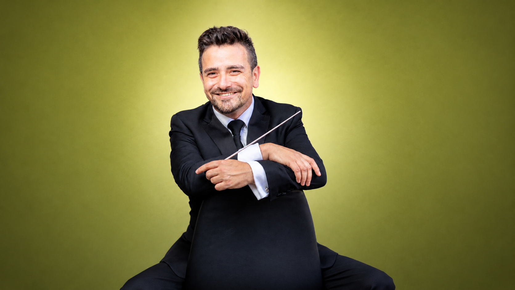 Andrés Lopera latino conductor seated in a professional portrait smiling with a green background.