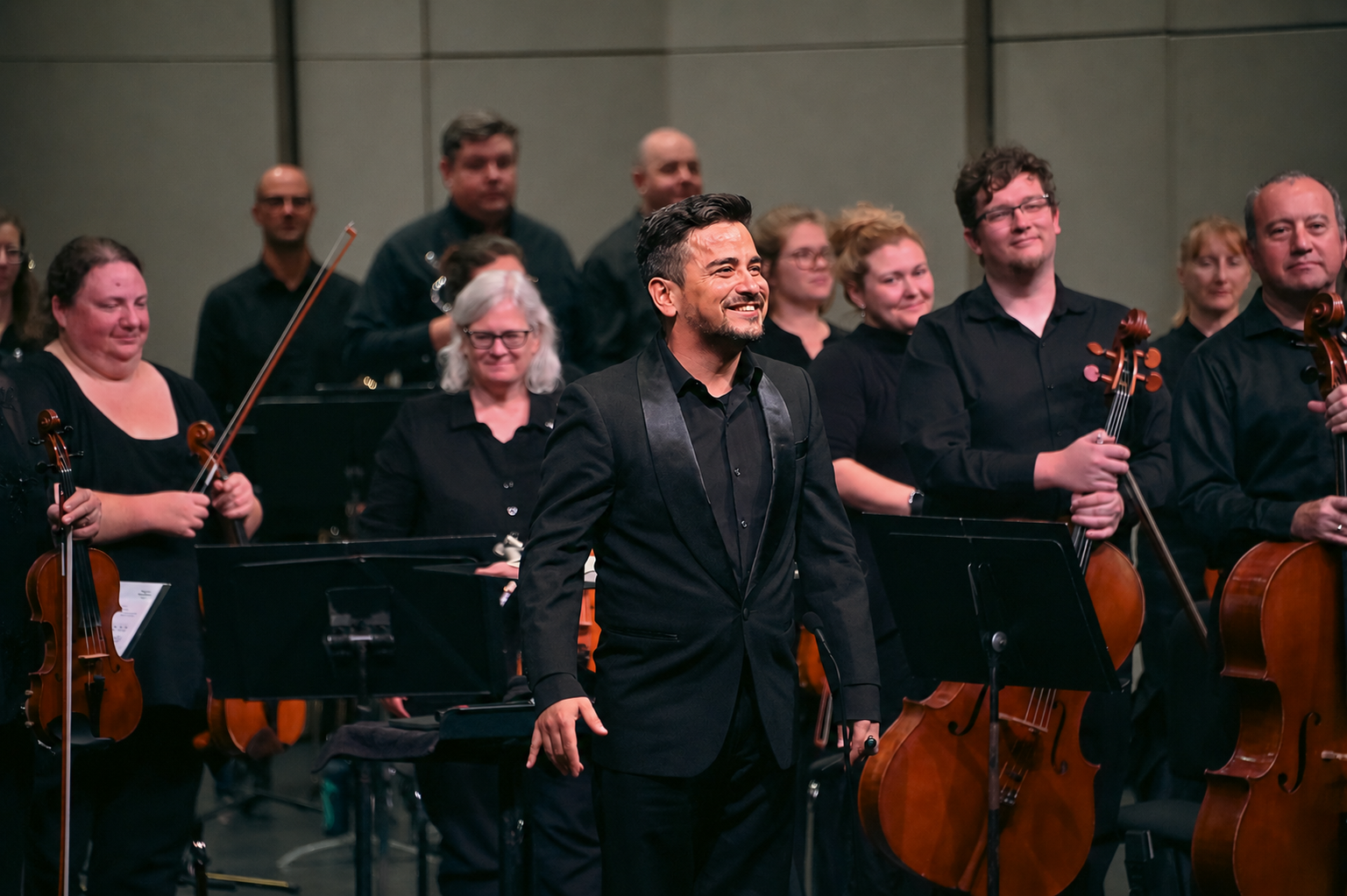 Andrés Lopera conducting a symphony orchestra during a live concert.. Orchestra members standing on stage after a performance, some holding violins and a Lopera in front taking a bow, smiling.