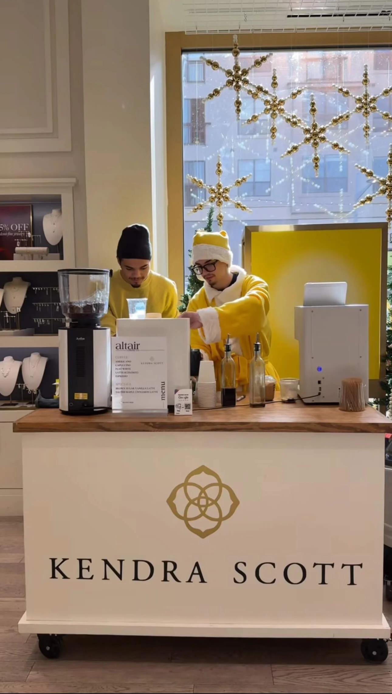 Two people behind a coffee cart counter with a Kendra Scott logo in Austin, Christmas decorations, large window with snowflakes, and jewelry displays in the background.