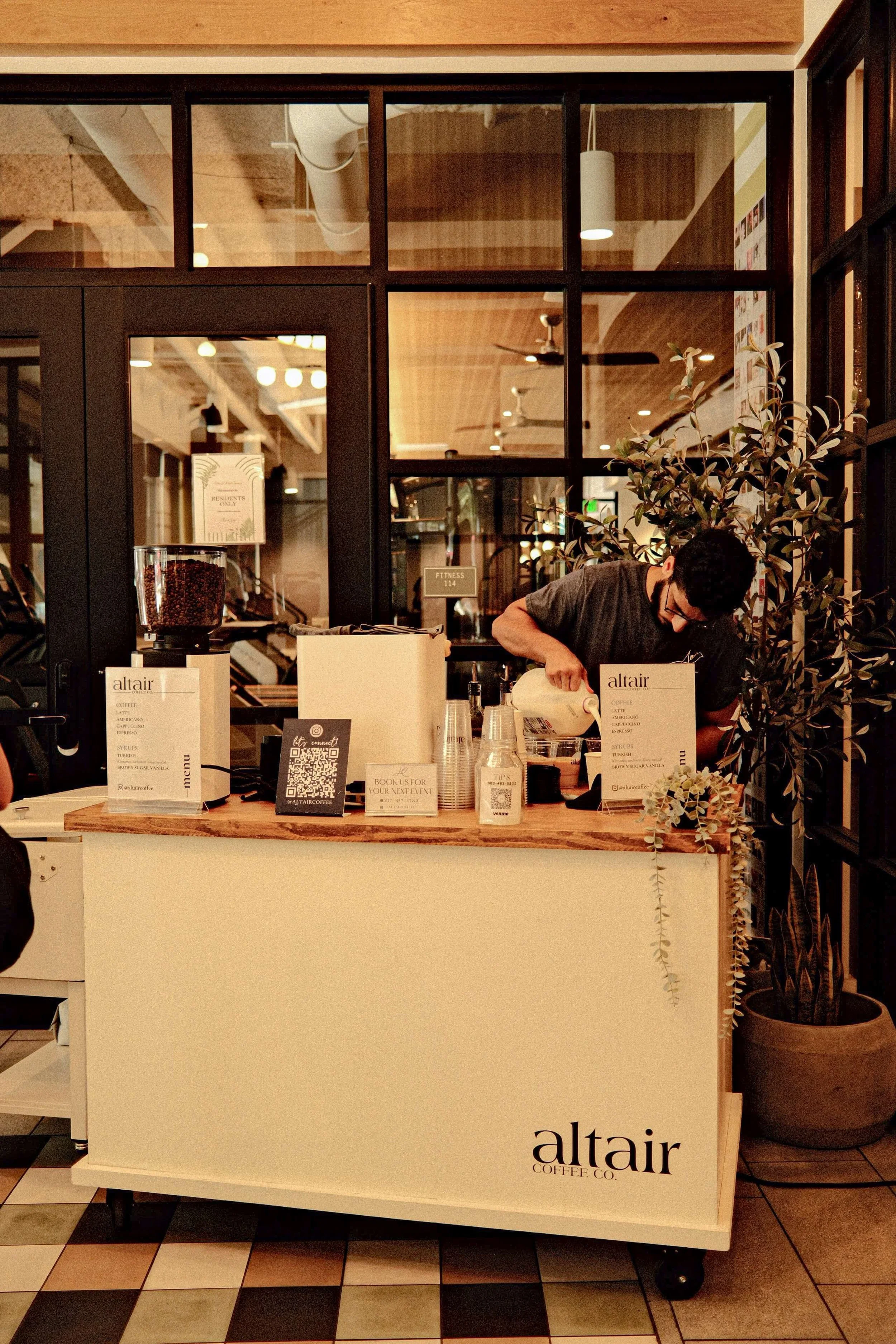 A coffee cart counter with a professional barista pouring milk, with signs displaying menu and QR code, and a large potted plant next to the counter, in a modern cafe setting.