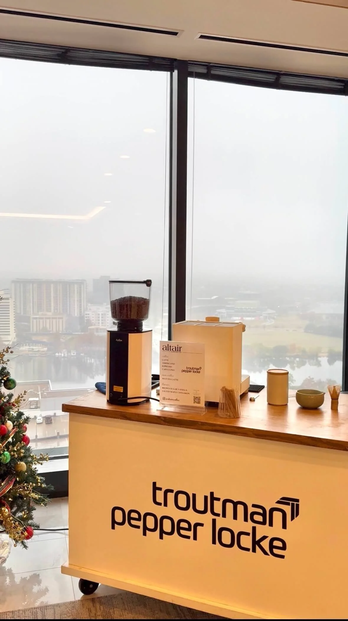 Coffee Cart station with coffee cart catering equipment, i.e coffee grinder, cups, and a menu sign, beside a decorated Christmas tree, with a large window overlooking a cityscape and water body in the background.