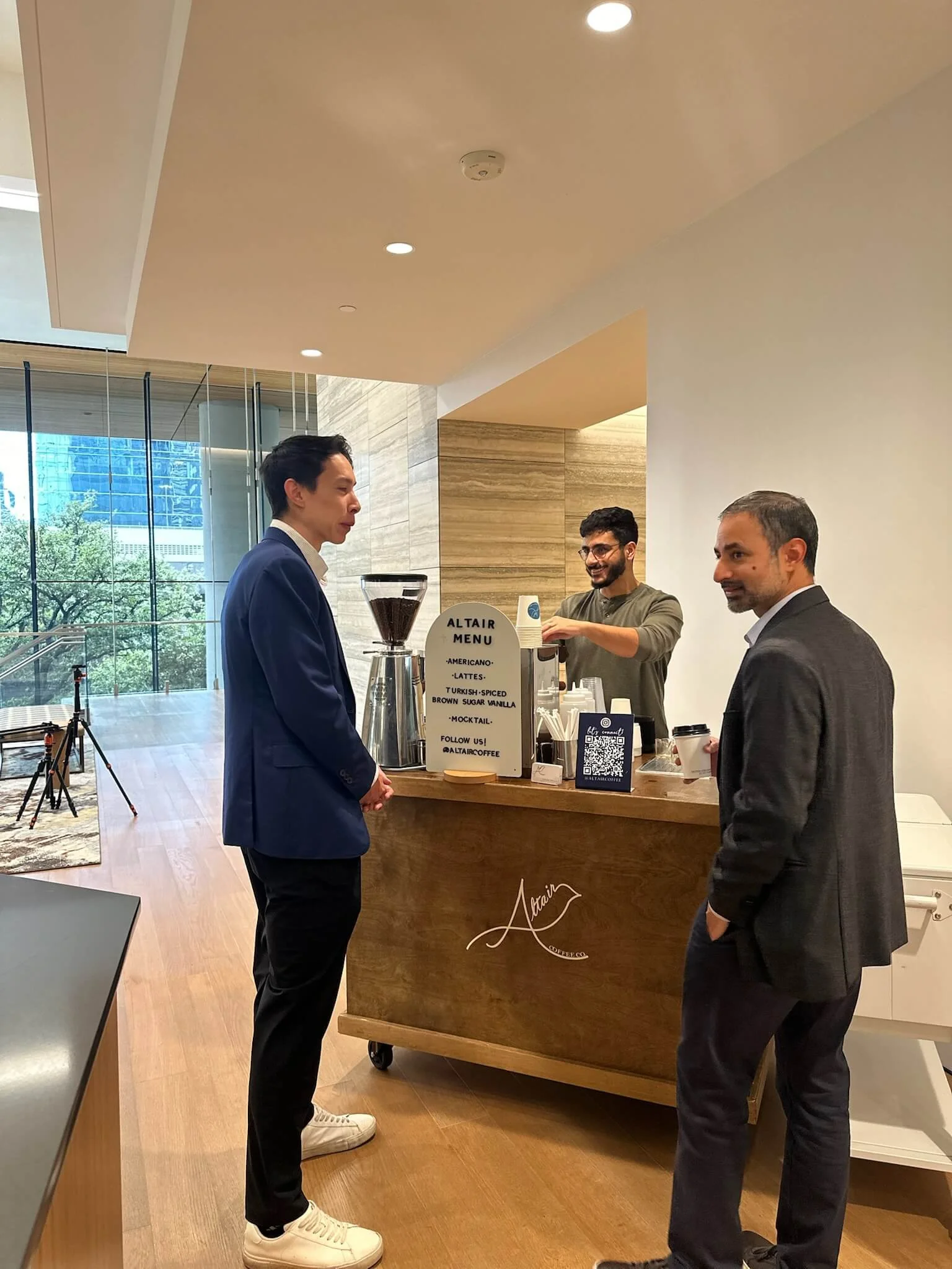 Two men in business attire ordering coffee at a coffee cart in a corporate office or hotel lobby with glass walls and a view of trees outside. The barista is smiling and preparing beverages behind the coffee bar.
