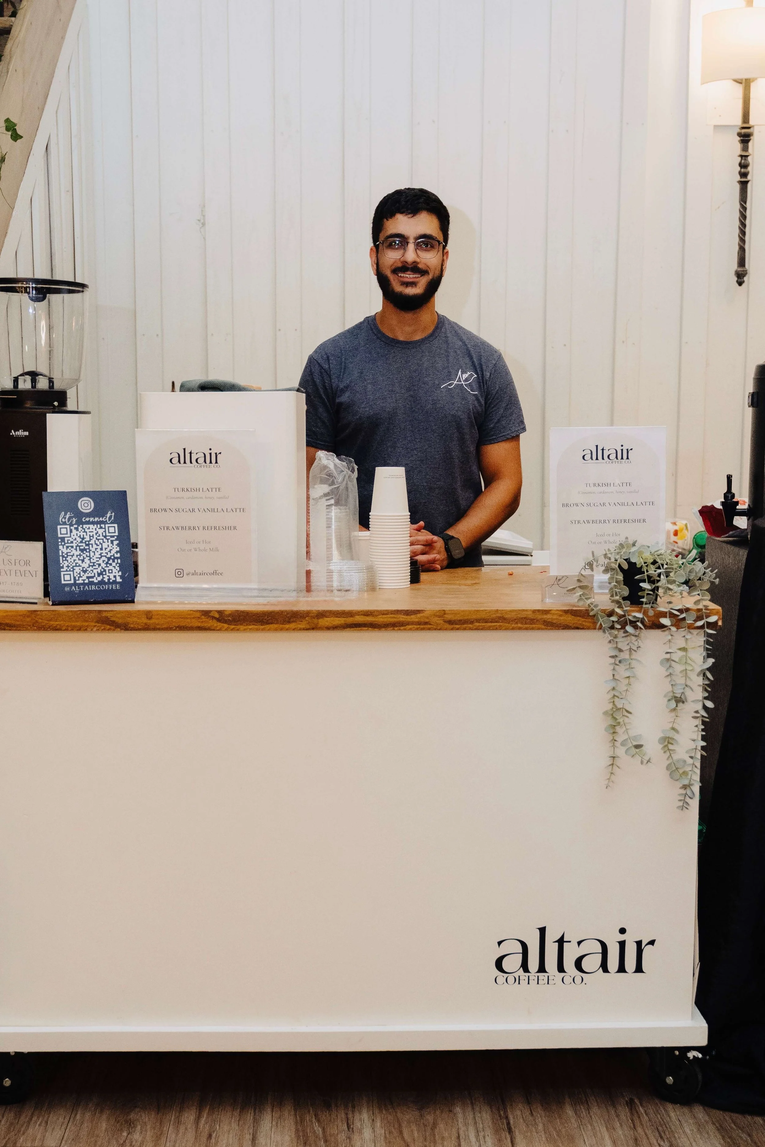 A smiling man standing behind a coffee cart counter with 'altair coffee co.' branding, serving coffee drinks, with signs displaying menu items like Turkish latte and strawberry refresher.